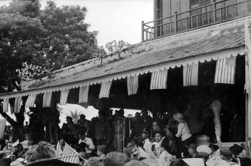 A crowd of people gathered under a roof overhang that is decorated with South Vietnamese and American flags.