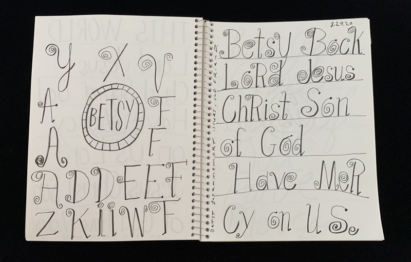 A spiral-bound notebook page featuring a variety of calligraphic and decorative lettering, including a circular design and a religious phrase.