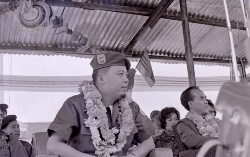 Nguyá»…n KhÃ¡nh is sitting at some event with a flower necklace around his neck.