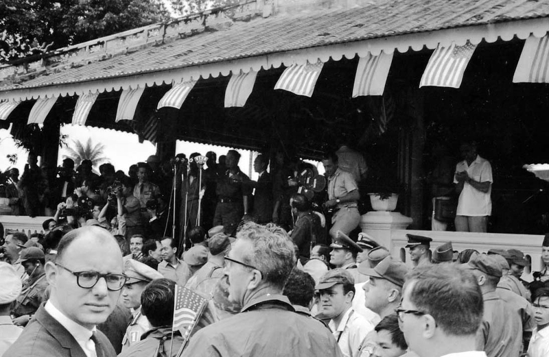 A crowd of people gathered under a roof overhang that is decorated with South Vietnamese and American flags. A Vietnamese man stands in front of microphones giving a speech.