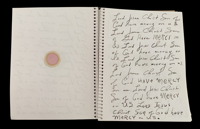 A handwritten page with repeated prayers and a small circular object on the left side.