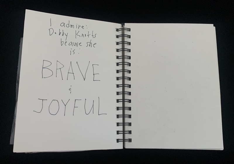 A handwritten note in a spiral-bound notebook lists admiration for Debby Knotts, highlighting her bravery and joyfulness.