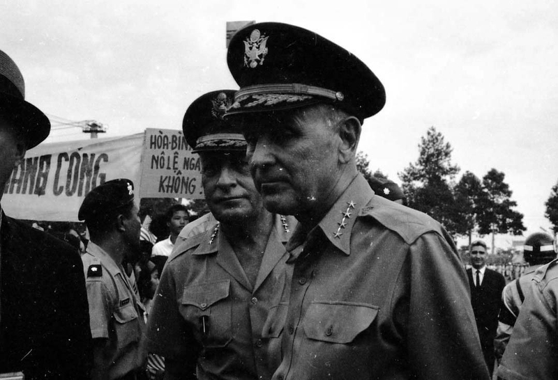 An American military officer that appears to be Maxwell D. Taylor in uniform with other military officials. There is a crowd with banners in the background.