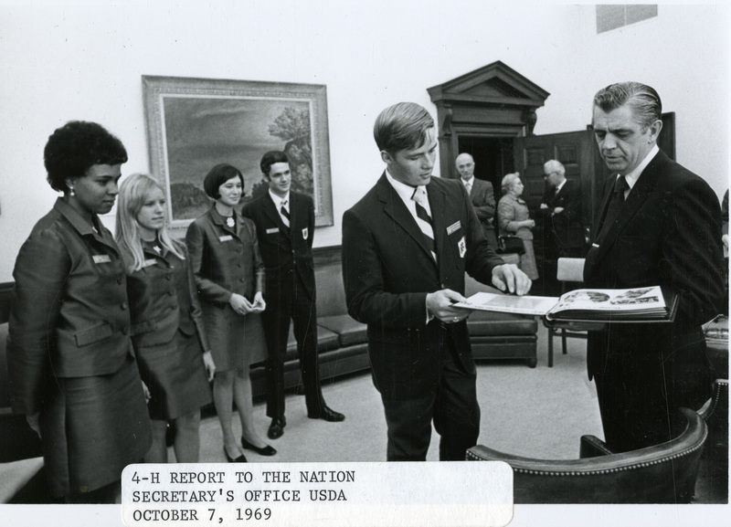 Original caption: "4-H report to the Nation Secretary's Office USDA October 7, 1969." Clifford Hardin looks at a scrapbook with a representative of 4-H, with four additional unidentified representatives looking on, an unidentified woman and two unidentified men stand in the office doorway.