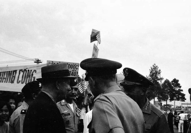 A group of American men are gathered in a group discussing something. Some are in military uniform and some are in civilian clothes. There is a crowd with banners in the background.