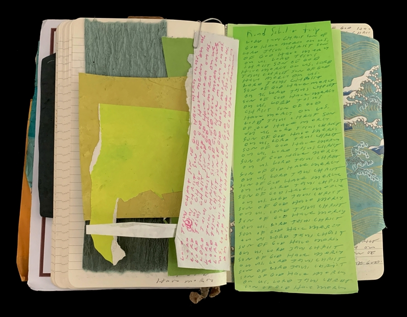 An open notebook with layered textured and colored papers, featuring handwritten text on green and white sheets.