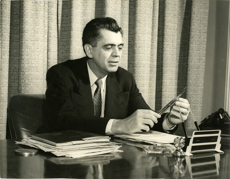 Clifford Hardin shown holding two pipes in front of three shelves of pipes. His Society of Innocents award hangs on the wall behind him.