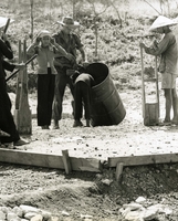 "'Ooops, not much water left.' John Murphy holds the water barrel, and old 55-gallon fuel drum - as one of his younger and smaller fellow workers 'gets to the bottom of things'. Water for the market project was carted by shoulder - Vietnamese style - by elderly women from a well half-a-mile away. Vietnamese construction methods were at first a bit strange to the American Seabees. Mixing concrete for instance, is done on the ground, at the spot where it will be used. No power mixer was used, only a wooden hoe, feet, and hands - the results are surprisingly good." Vietnamese workers pouring concrete in a build site. There appears to be an American soldier and a Vietnamese child also working.