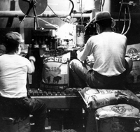 "Korea-Negs 181-3(1)-182-3(1) Fertilizer Plant-Chungyu- USAID 7" Two men sit facing away from the camera filling bags with fertilizer on an assembly line. The machine they are using appears to be sealing the bags. Their faces are not visible, and they appear to be wearing protective masks. From scrapbook page 23.