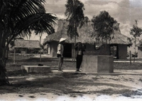 Two villagers stand by a concrete well.  There are thatched houses in the background.