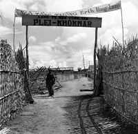 "Montagnards - Showplace Strat. Ham - 8-21-62 - In MTs - Faces - Blockhouses" A Vietnamese soldier is standing under a banner surrounded by wooden fences on either side.