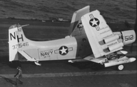 "7th Fleet" A Naval plane on an aircraft carrier. The wings of the plane are detached and folded. An American man in uniform can be seen nearby, perhaps giving directions.