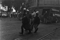 "Embassy Bombing" Three firemen carry a body on a blanket from the wreckage. Behind them, another fireman runs a hose from the back of the firetruck. People cluster in the left corner.
