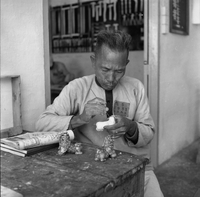 "Ai Lam - HK*" A man is carving small Chinese-style lion statues.  His uniform suggests he is a prisoner.