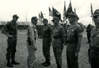 "Cu Chi, Vietnam - Major General Fred C. Weyand (left), 25th Infantry Division commander, welcomes General John K. Waters (second from left), commander-in-chief, U.S. Army Pacific, during color ceremonies at the Cu Chi base camp Saturday, July 23. In the background are members of the Division's staff, along with color bearers." An American commander stands and talks to a row of soldiers. More soldiers with flags stand in formation in the background. They appear to be in a military camp.