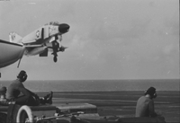 "7th Fleet" A Naval plane taking off from an aircraft carrier. Three American men wearing headsets sit and watch it fly.