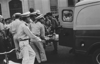 "Embassy Bombing" A crowd of people in the street. Men in uniform lift a man on a stretcher into the back of an ambulance.
