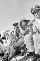 "Unidentified", A goup of spectators watch an event from some bleachers. The two men siting in the middle of the photo appear to be Nguyễn Khánh (holding binoculars) and possibly Nguyễn Cao Kỳ (smoking). Other spectators are Vietnamese men in civilian clothes, Western men and women, and a Catholic priest.
