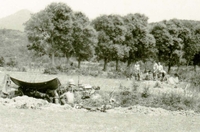 Two American soldiers sit in a trench shaded by a tarp and other soldiers are seen in the background