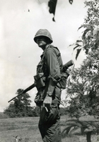 "Bien Hoa, Vietnam (IO) - Army Sgt. Richard J. Manley stops a moment to check the area during operation 'Plum Bob.' Sgt. Manley is a member of Co. 'C', 16th Inf., 1st Div." An American soldier stands in a field holding a gun and a cigar.