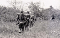 Several American soldiers walking through a field in a line.