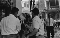 "Embassy Bombing" Two men take a statement from a man with a bandaged head. A second man holds the bandaged man up with one hand and holds a hankerchief to his mouth with the other. Behind them stands a ruined building and more people.