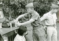 An American soldier named Hibbs is digging through a box of band aids to treat an injured woman sitting on the bumper of a Jeep. A second soldier is in the background.