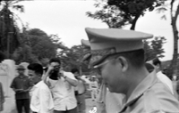 "Unidentified", General Nguyễn Khánh walks by in military uniform. The background is blurry, but there are several men taking photos in the background.