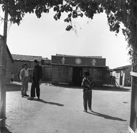 "Macau" A young child is facing some wooden buildings as two men are conversing to her left.