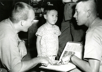 A young Vietnamese child is looking at two American soldiers as they are pointing at photos in a photo album.