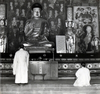 "Korea-Negs 183-1(2)-184-1(2) 6.Kyongju" Two people face away from the camera in a Buddhist temple. One sits on the floor and appears to be praying. The other, possibly a monk, stands in fron t of an altar with a large Budha statue standing over him. There are murals on the wall abd other statues on a platform. From scrapbook