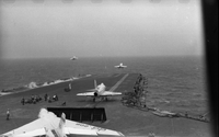 Men working on an aircraft carrier in open water. Two Naval planes are still on the runway. Two have just taken off.