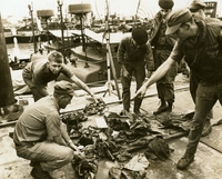 "Enemy clothing, personal equipment and Russian-made sighting device are inspected by U.S. Navy river patrol boat crewmen who captured them early this morning. Crewmen of the PBE-753 and PBR-754, based at Nha Be, eight miles South of Saigon, killed at least 15 enemy troops believed to have been involved in the rocket attack on U.S. Army airfield at Vung Tau, 35 miles southeast of Saigon." A group of American soldiers stand around a pile of clothing and equipment.