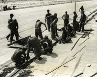 "Seabees from Seventy-One's Charley Company are strung out along the center seam of Chu Lai Main Runway, as they repair the seam with specially prepared materials. The 2-miles seam had deteriorated and was endangering fast landing aircraft. The Seabees were called on to cut out the broken concrete and replace it with a special epoxy glue and specially mixed concrete. The concrete, requiring an extremely short "set-up" time was mixed close to the seam and quickly put into place by the 25 Seabees that worked on the project 24-hours a day. The project was completed four days ahead of schedule and won a pat on the back of Commodore Turner, Commanding Officer of the third Naval Construction Regiment." A group of American soldiers pour concrete in a narrow ditch. They appear to be on an air field runway.