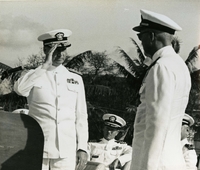 "Change of Command - Rear Admiral Kenneth L. Veth, USN (left) salutes Rear Admiral Norvell G. Ward, USN, at a ceremony 27 April 1967 at which RADM Ward as COMNAFORV. The ceremony was held aboard the USS Garrett County (LST 786) docked at Saigon, South Vietnam." An American man in formal military uniform salutes another uniformed man.  Palm trees and more uniformed men sit in the background.