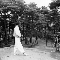 "Korea-Negs 182-3(2)-183-1(1) 6.Baggy Pants" A Korean man dressed in all white walks on a dirt path through a forest. In the far right of the frame there is a market stall. From scrapbook