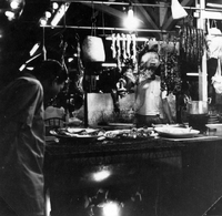 "Sarawak." An outdoor night market stall selling barbequed meats. A man stands behind the barbeque with plates of meat and skewered meat hangs above him. A customer looks at the meat on sale. From scrapbook page 13.
