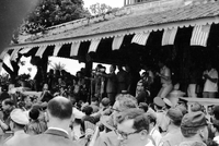 A crowd of people gathered under a roof overhang that is decorated with South Vietnamese and American flags. A Vietnamese man stands in front of microphones giving a speech.