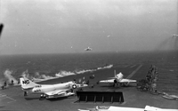 Men working on an aircraft carrier in open water. Three Naval planes are still on the runway. Two have just taken off.