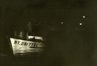 "Night Work - A Vietnamese junk is picked up at night by a search light from the U.S. Navy Patrol Gunboat USS Gallup (PG 85). Boarding parties must be prepared 24 hours a day to conduct searches for contraband goods the enemy attempts to infiltrate to Viet Cong ashore in South Vietnam." A dark photograph of a boat at night.