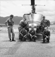 A group of American soldier pose for a photo in front of helicopter. One has his head bowed and another is writing something on the helicopter.  A monkey on a leash sits at one soldier's feet.