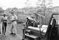 An American soldier named Hibbs is digging through a medical bag sitting on the hood of a Jeep. A second soldier is in the background.