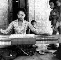 Original title: "Indonesian Arts and Crafts Negs [Negatives]." An Indonesian woman sits behind a loom and weaves a piece of cloth. She is watched by three young children. From a scrapbook page 10.
