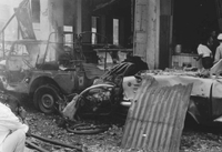 "Embassy Bombing" Two cars have collided in the steaming rubble. Two men enter a ruined building. A man crouches in the debri-ridden street to take a photograph.