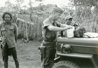 An American soldier named Hibbs is digging through a medical bag sitting on the hood of a Jeep. Several villagers are in the background.