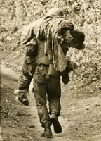American soldier is seen carrying a man on his back down some type of path. The soldiers back is to the camera.