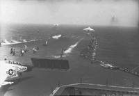 Men working on an aircraft carrier in open water. One Naval plane is still on the runway. Three have just taken off.