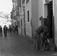 "Macau" A man is leaning against his bike speaking to someone through a doorway, as several others walk down the street in the background.
