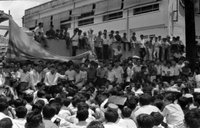 "Unidentified", a large crowd of young Vietnamese men, sit outside watching a man give a speech . A large South Vietnamese flag hangs behind him. In the background, more men stand behind the speaker and sit on a wall . This is likely a part of the anti-Diệm student protests.