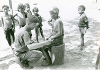 An American soldier named Hibbs examining an elderly man's hand. Villagers watch in the background.